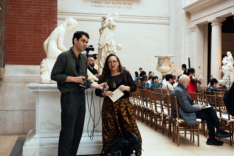 A tall man and a shorter woman are standing in front of a marble statue of a person sitting. Behind them are several people sitting in rows of chairs.