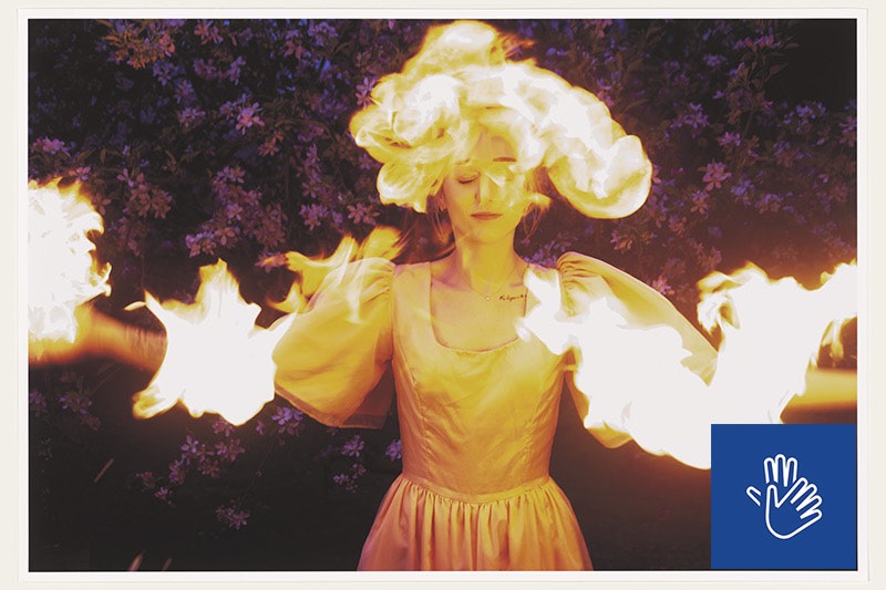 A woman wearing a silk gown with puffy sleeves stands with arms outstretched in front of a background of purple flowers. Flames engulf her head and arms while she smiles serenely with eyes closed.