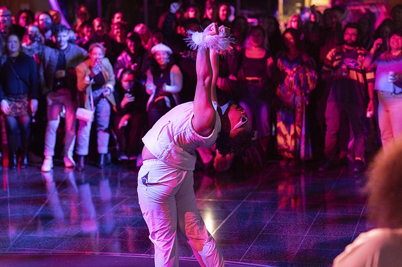A person in a white top and trousers, with white fur around their wrists is performing in front of a crowd under red and blue lights.