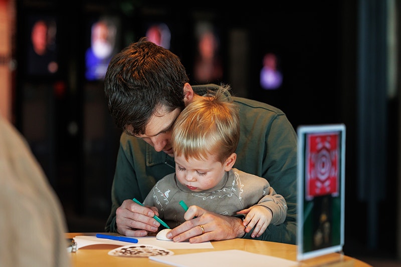 An adult has a toddler on their knee and they are both colouring in a piece of paper on the table in front of them.