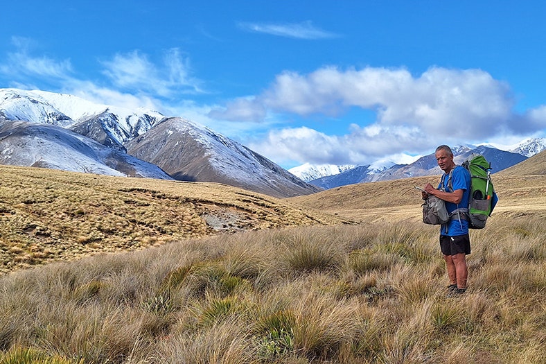 A man in hiking gear and carrying a pack is making notes in a notebook. He is standing in a tussock field that has mountains in the background that have a light covering of snow.