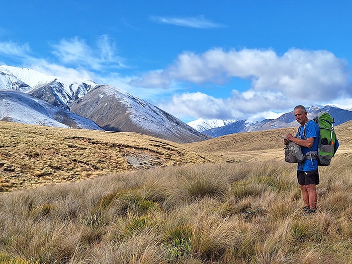 Colin Miskelly counting pipits below the snow-capped peaks of the Hakatere Range. Photo by Gordon Miskelly and Colin Miskelly A man in hiking gear and carrying a pack is making notes in a notebook. He is standing in a tussock field that has mountains in the background that have a light covering of snow.