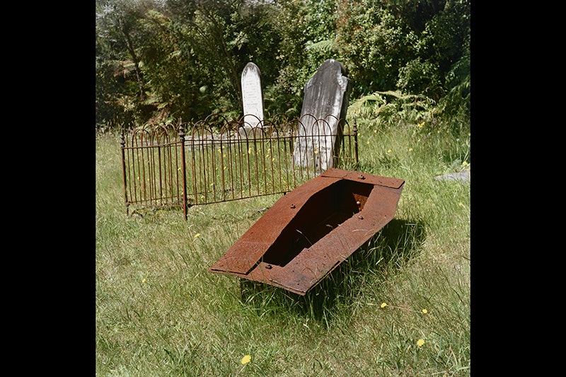 A rusted metal box in the shape of a coffin is sitting on top of grass in a cemetery.