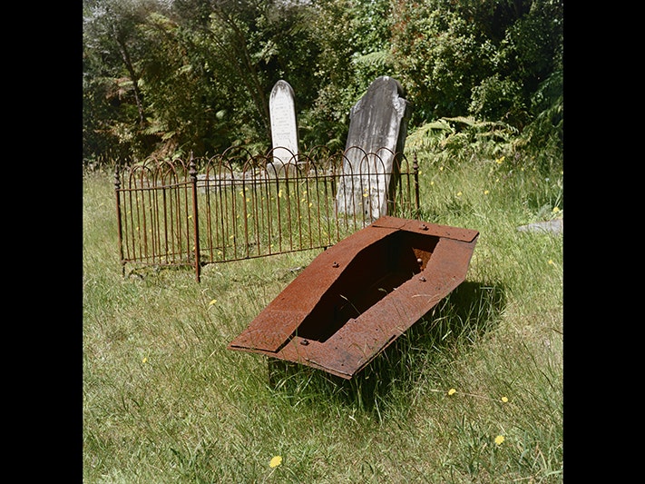Caroline McQuarrie, Metal child-size coffin, Stafford cemetery, December 2011. From the series: No Town. Purchased 2024. Te Papa (2024-0030-7) A rusted metal box in the shape of a coffin is sitting on top of grass in a cemetery.