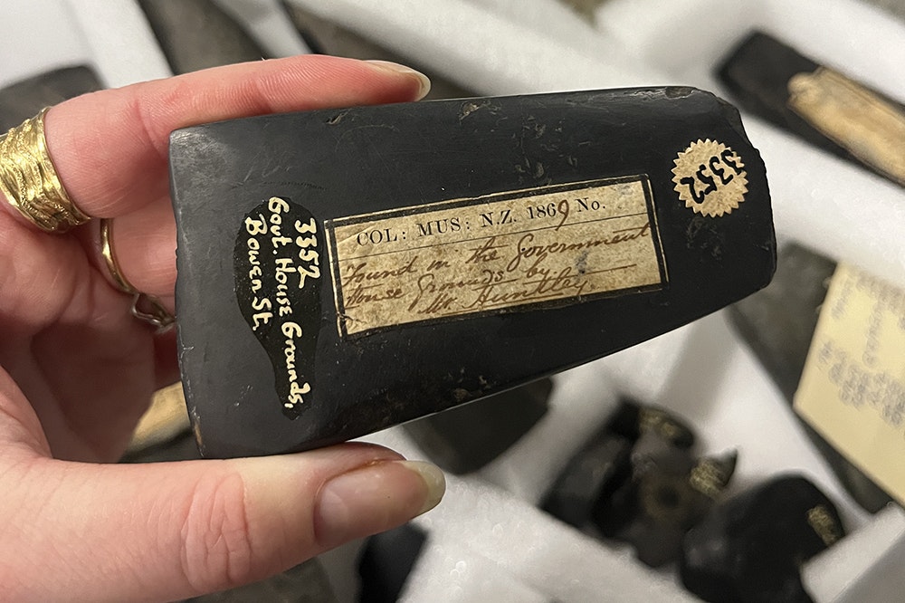 A hand is holding a stone adze with old museum stickers and handwritten labels on it.
