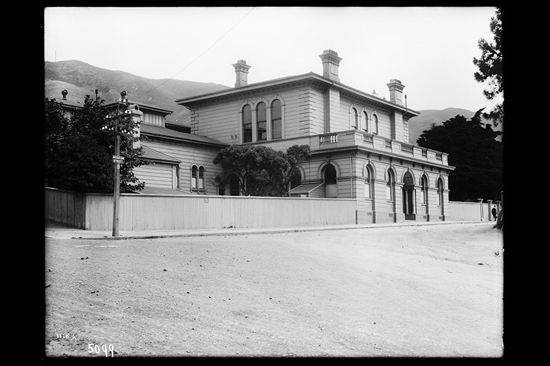 A black and white photo of a wooden building with three chimneys viewable, and a wide street in front of it.