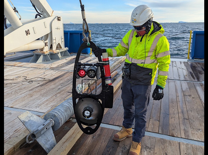 Lure deploy. Photo by Thom Linley. Te Papa (257271) A person in high vis gear is holding piece of scientific equiment on a rope on a boat.
