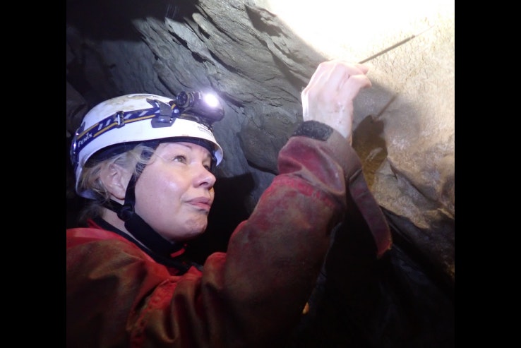 A woman with a headtorch on her helmet is poking at something in a cave.